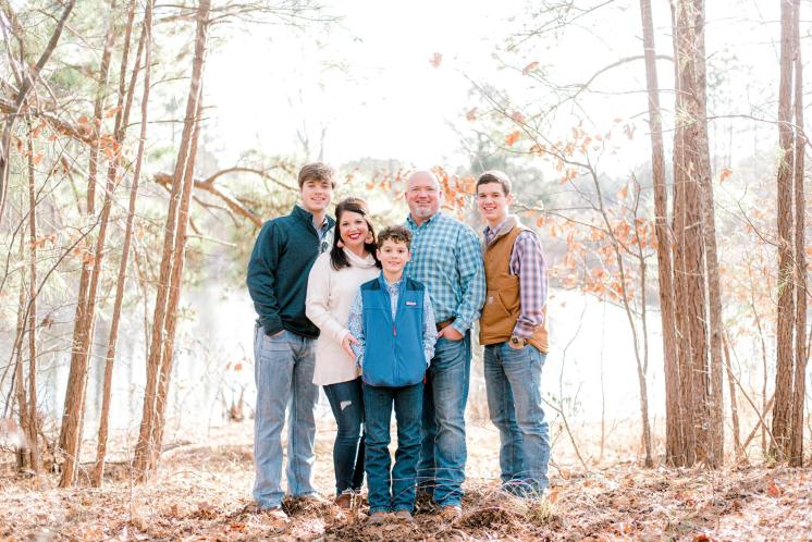 A group of five people standing together on a forest path with trees and a lake in the background.