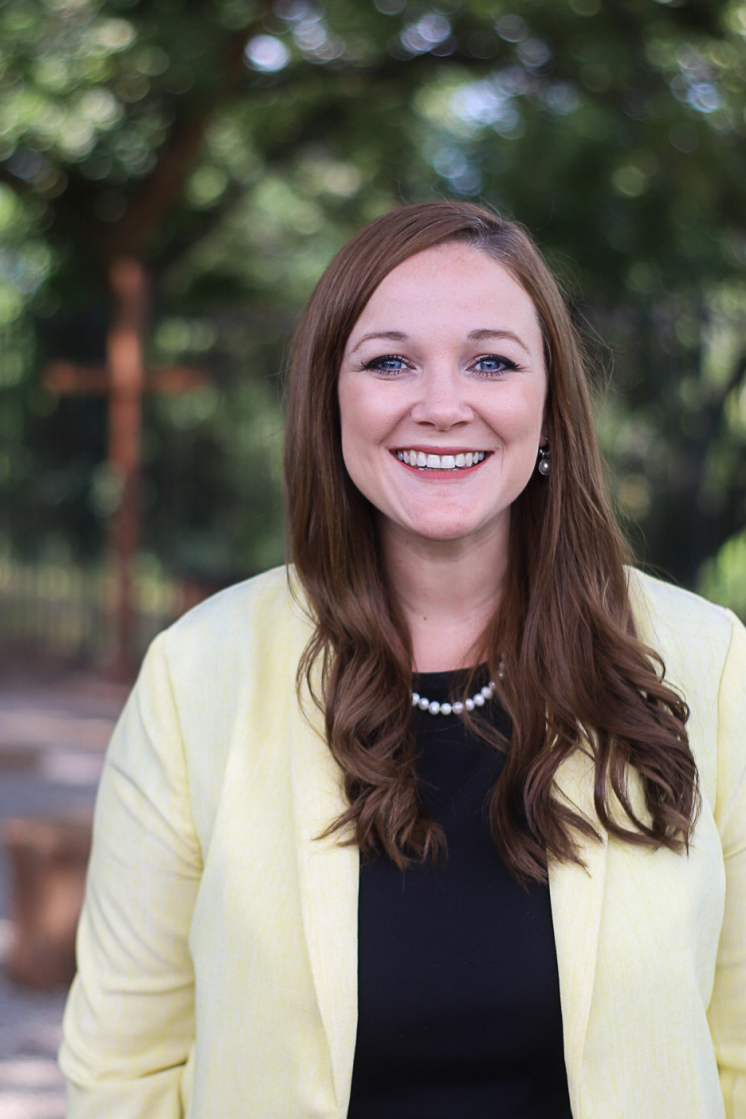 Headshot of a woman in a blazer and black shirt smiling in front of green trees.
