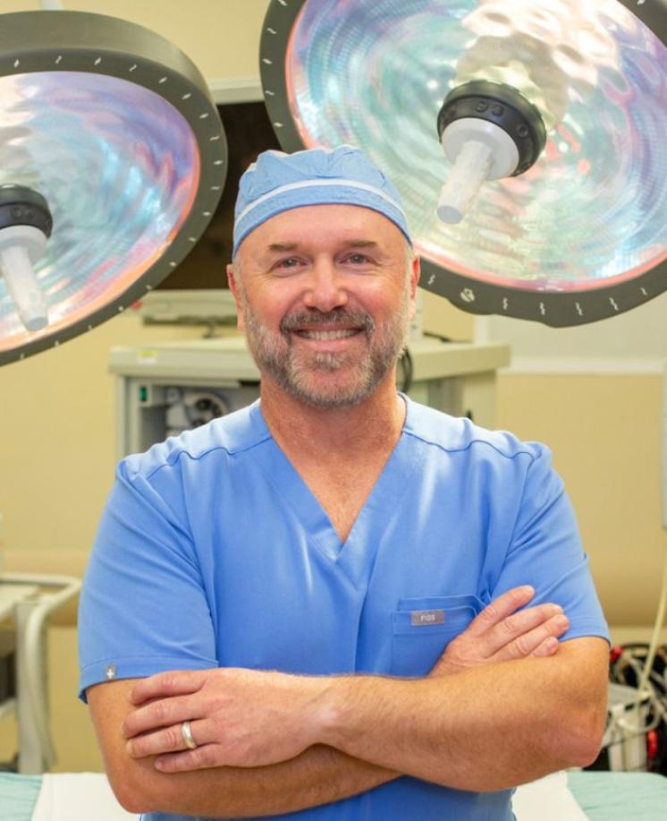 A man in light blue scrubs and scrub hat stands in an operating room with lights behind his head.