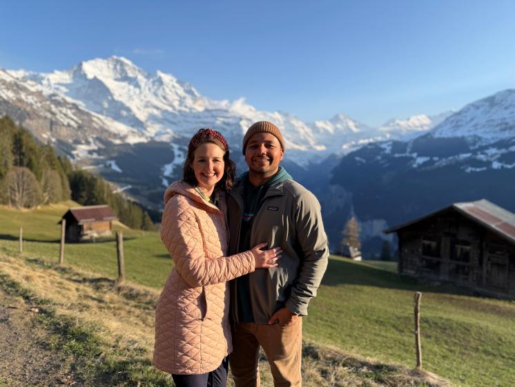 2 people standing in a field with snow capped mountains behind them