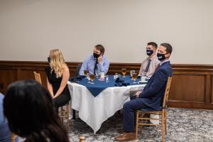 People seated around a table set for a formal event, wearing dark face masks.