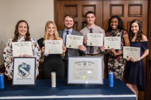 Group of six people holding certificates behind a table displaying framed documents and candles.