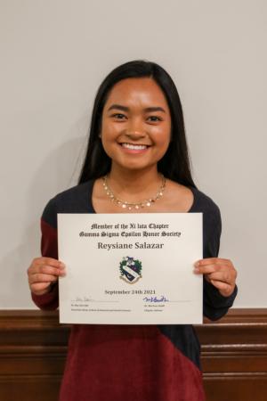 Individual smiling and holding a certificate in front of a light-colored wall.