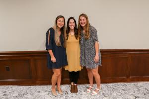 Three women standing and smiling indoors against a wall with wooden paneling.
