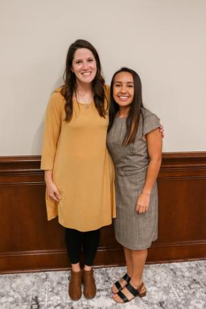 Two women standing and smiling indoors against a wall with wooden paneling.