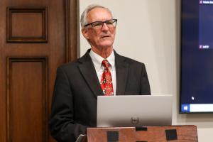 An man in a suit and tie stands at a podium with a laptop, with a wooden door and large screen in the background.