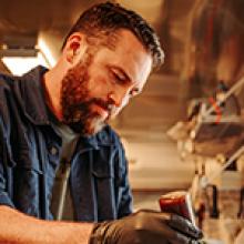 Man with a beard working in a workshop or kitchen, wearing gloves and a denim shirt.