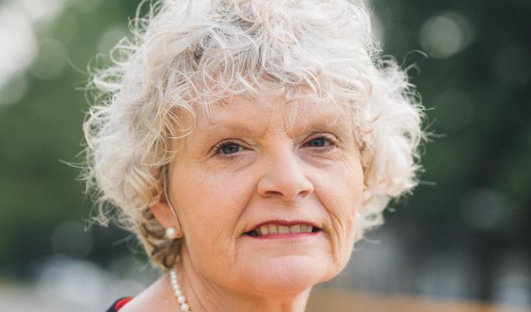 Older woman with curly white hair, wearing a red blouse and pearl necklace, against a blurred outdoor background.