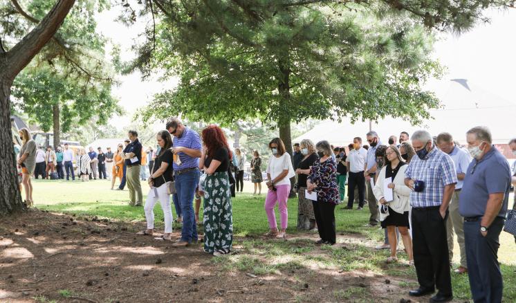 A group of people stands on a grassy area under trees, some wearing casual and some in semi-formal attire.
