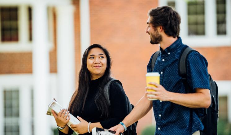 Two students walking and talking in front of a brick building on a college campus.