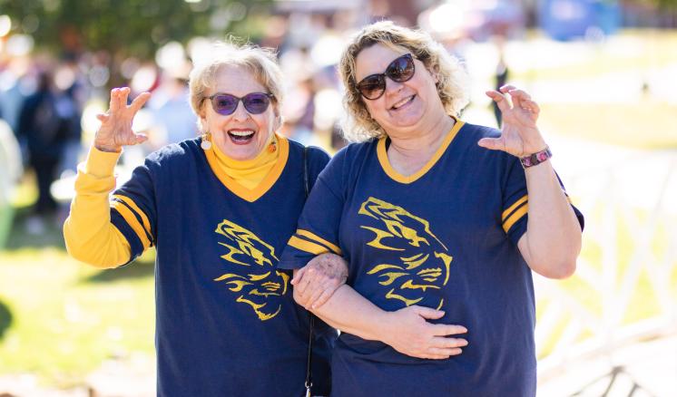 "Two people outdoors wearing matching blue shirts with yellow graphics, both smiling and making hand gestures."