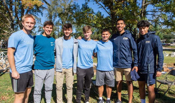 A group of seven young men standing outdoors, wearing casual clothing in blue tones, with trees and grass in the background.