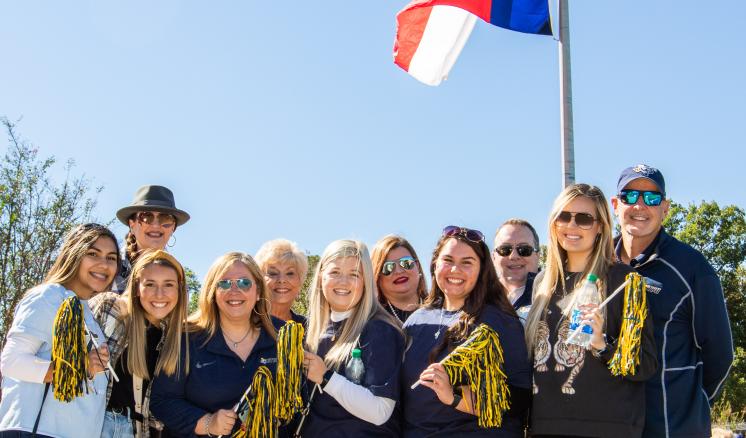 A group of ten people standing together outdoors, holding gold pom-poms with flags in the background.