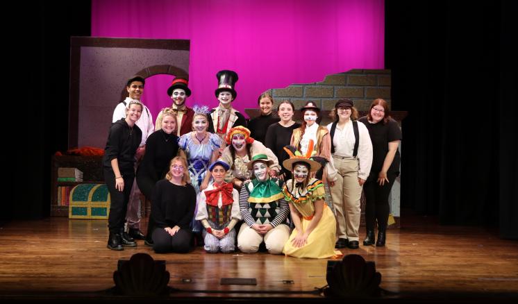 A group of performers in colorful costumes on a stage with a pink-lit background.