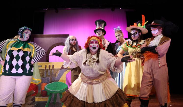 A group of people in colorful theatrical costumes posing on a stage with a pink backdrop.