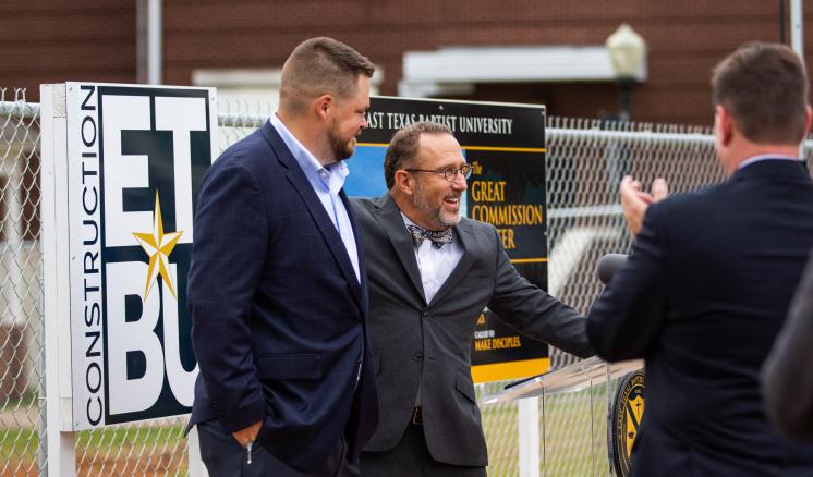 Three men in suits talking in front of a fence with signs.