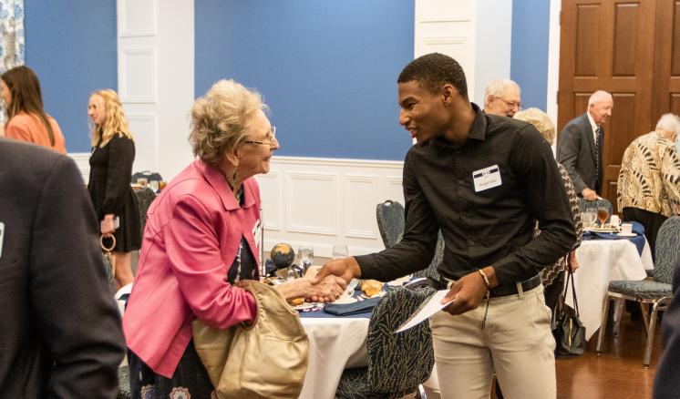 An elderly woman and young man shaking hands at a formal gathering in a blue-walled room.