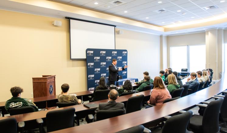 A conference room with attendees seated at a U-shaped table, listening to a presenter at the front.