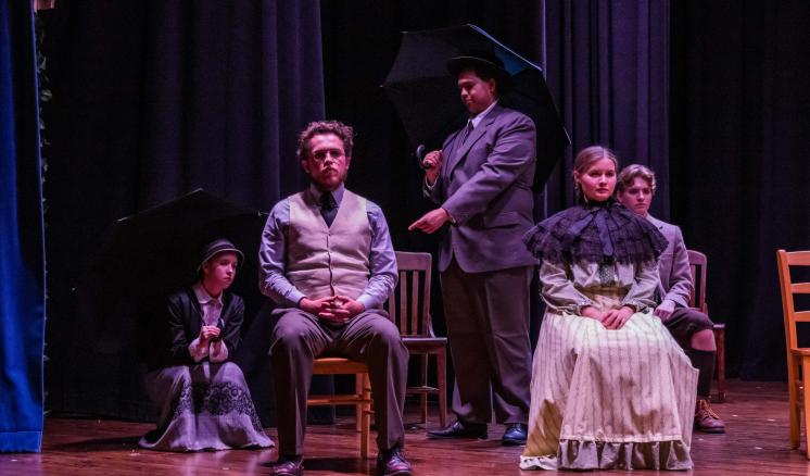 A theatrical scene with five people in period costumes, some holding black umbrellas, seated on wooden chairs on a stage.