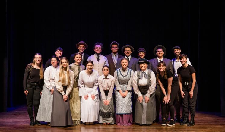 A group of seventeen people in period costumes pose onstage.