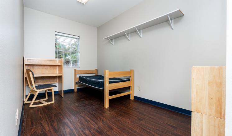 A small room with a single bed, desk, and chair, featuring white walls and dark wood flooring.