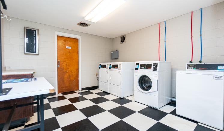 A laundry room with checkered black and white flooring, three washing machines, and utility pipes on the wall.
