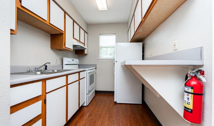 Narrow kitchen with white cabinets, a gray countertop, a stove, a refrigerator, and a fire extinguisher on the wall.