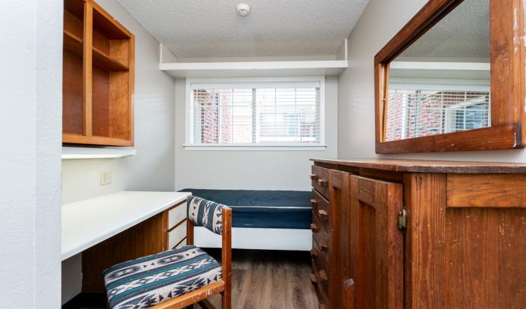 Small dormitory room with a bed, desk, chair, bookshelf, and dresser.