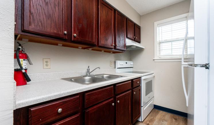Compact kitchen with dark wooden cabinets, white countertops, stainless steel sink, white stove, and a window with blinds.