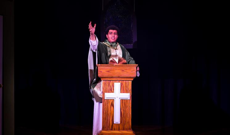 Person in vestments behind a lectern with a cross, on a dark stage.