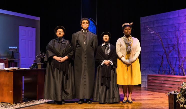 Four people in historical costumes on stage, with a desk to the left and brick wall backdrop.