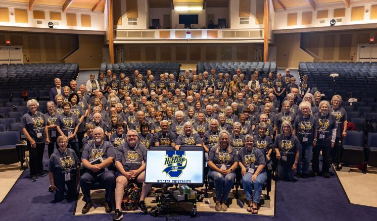 Large group of people in an auditorium wearing matching grey t-shirts, with a screen in front displaying a logo.