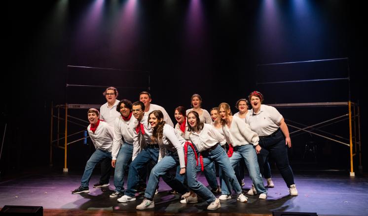 A group of performers in white shirts and blue jeans energetically pose on a stage with colorful lighting.