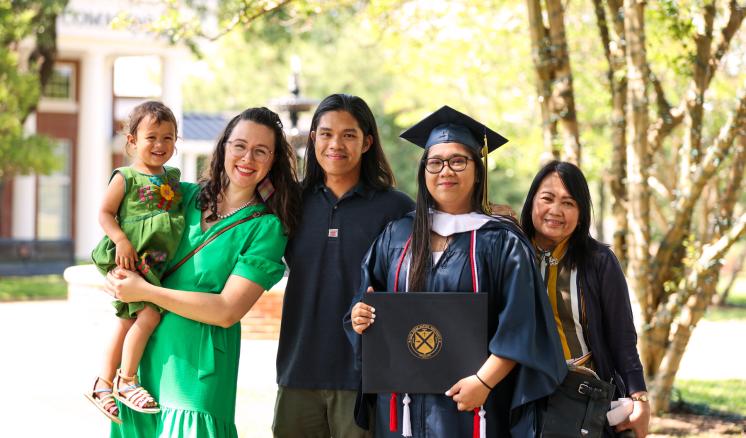 A group of five people, including a graduate in cap and gown, posing outdoors with trees and a building in the background.