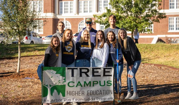  A group of nine people stand around a sign that reads "TREE CAMPUS Higher Education Arbor Day Foundation" with a brick building in the background.