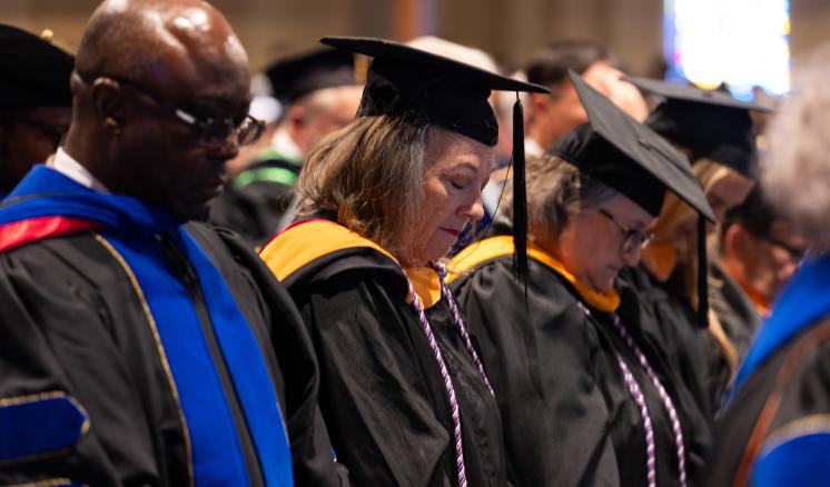 A group of faculty members, dressed in black academic regalia with colorful hoods, bow their heads in prayer or reflection during ETBU's fall 2024 Convocation chapel