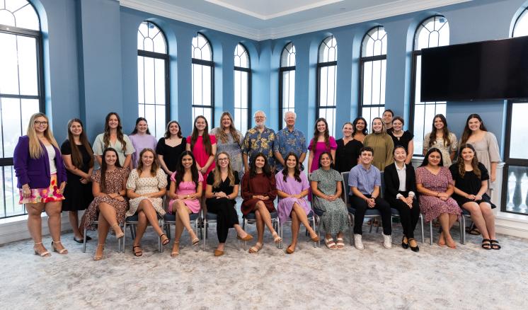 Group photo of a diverse team of individuals at East Texas Baptist University, posed in a bright, spacious room with large arched windows. The group consists of men and women, dressed in a variety of professional and semi-casual attire, seated and standing in two rows.