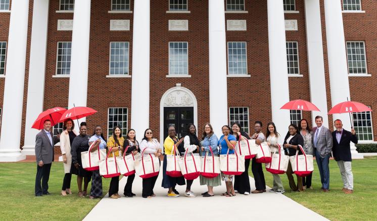 Group of people holding tote bags and red umbrellas, standing in front of a large brick building with white columns and windows.