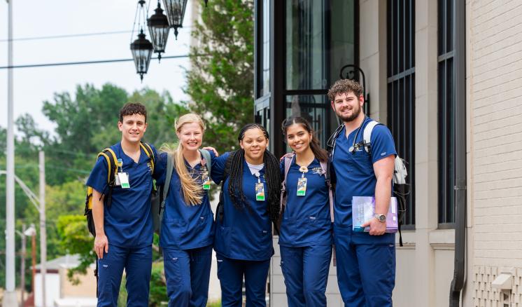 Five students stand side by side outside of building in nursing school scrubs, some holding textbooks and carrying backpacks