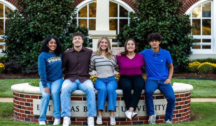 Five college students sit on a circular raised flower bed surrounding ETBU's Light on the Hill