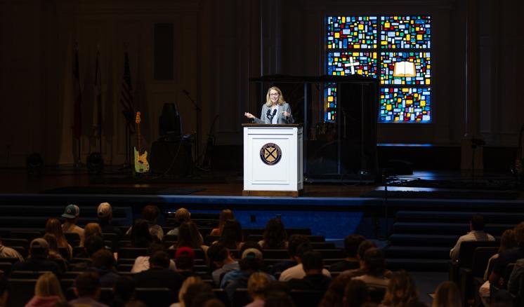 A woman stood on a stage in formal clothing addressing a congregation with a podium in front of her
