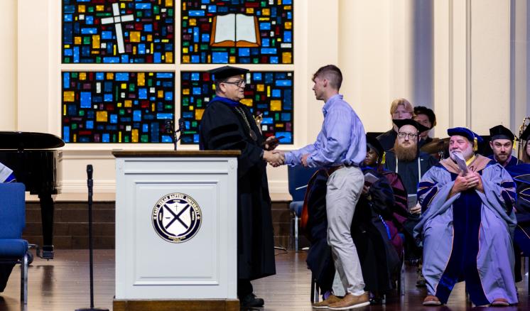 A man receiving an award on a stagr behind a podium while faculty in academic regalia sit behind