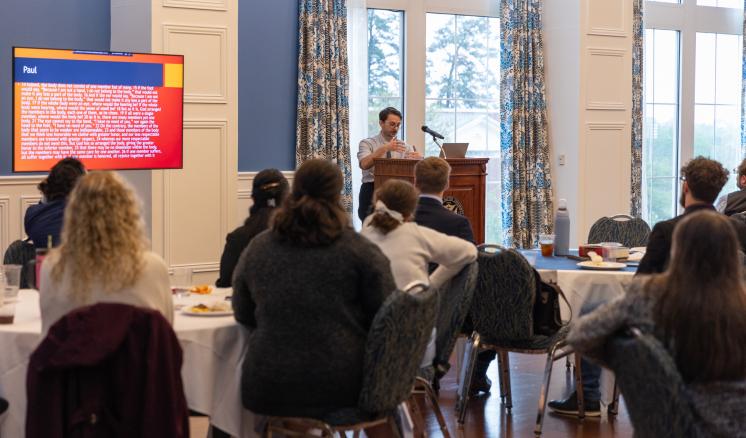 A group of people listening to a speaker talking behind a podium in a room with natural light from bright windows