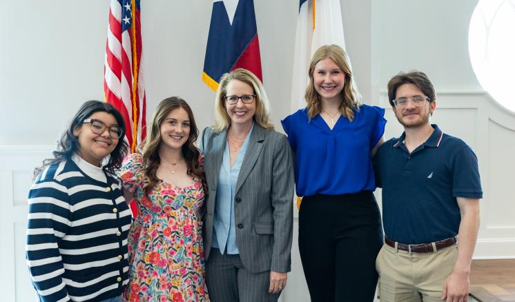 A group of 5 people stood in front of flags in a well lit room smiling