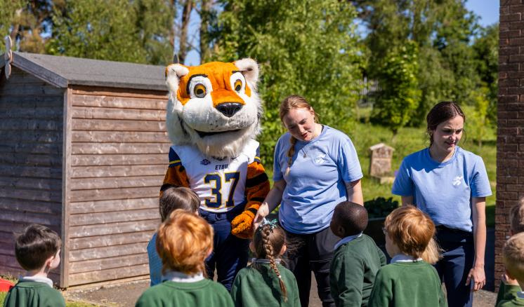 A costumed tiger mascot with the number 37, flanked by two adults, engages with children in green tops outdoors near a shed and trees.