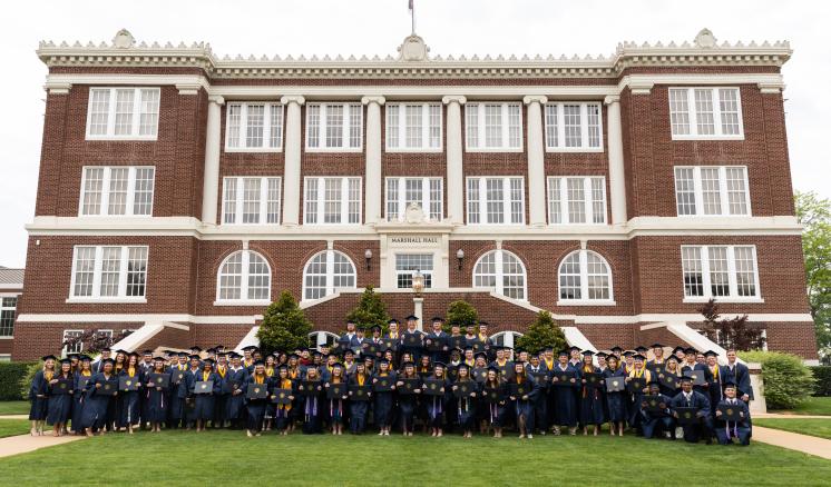 A group of graduates in gowns and caps pose in front of a large brick building with white columns.
