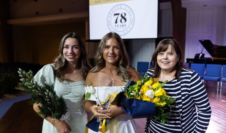 3 women posing together with flowers in their hands and smiling expressions