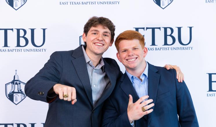 2 guys in front of an ETBU backdrop showing off their rings