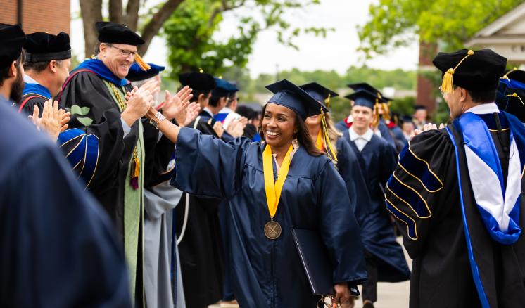A graduate in a cap and gown high-fives faculty members during an outdoor ceremony.