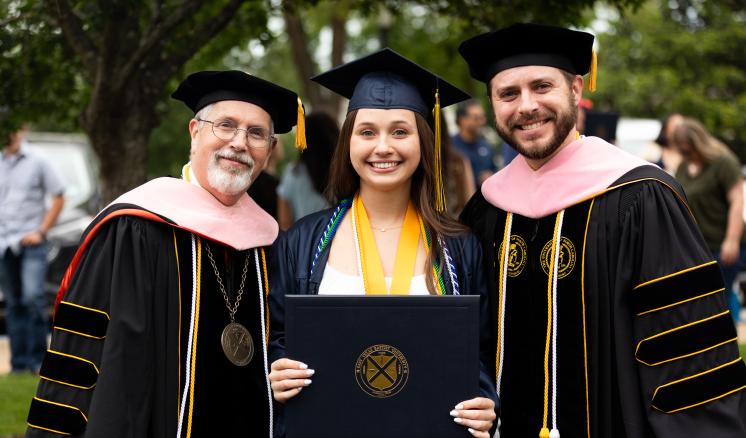 A graduate holding a diploma cover stands between two faculty members in academic regalia at an outdoor ceremony.